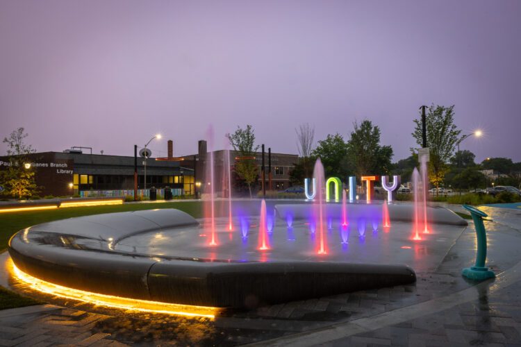 Splashpad in evening, lit up red and purple with colorful "Unity" letters behind.