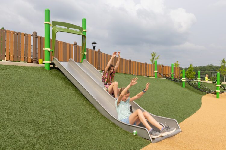 2 girls going down tan slides, side by side, with hands in air.
