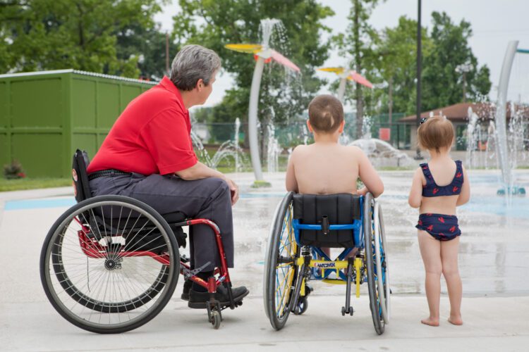 Parent in wheelchair next to boy in wheelchair and girl standing looking at water jets.