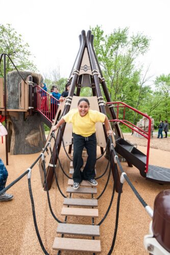 A girl walking across a bridge connected to a custom teepee play structure at the Indian Creek Playground in Berwyn Heights, MD, installed by Sparks at Play using LSI equipment.