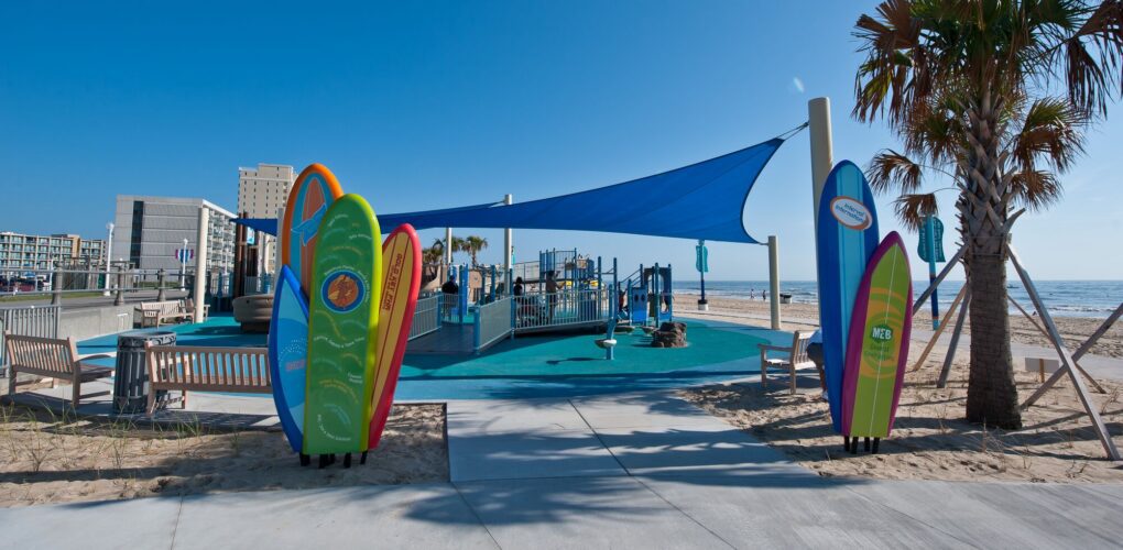 Entrance to JT's Grommet Island Beach Park Playground for EveryBODY in Virginia Beach, VA, featuring custom surfboard decorations and LSI SkyWays® shade structure, installed by Sparks at Play.
