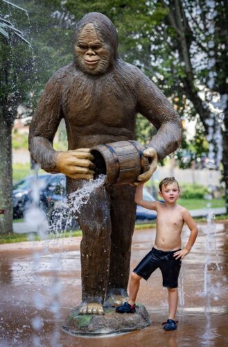 A little boy standing next to the custom GFRC Norton Wood Booger statue at the City of Norton splash pad in Norton, Virginia, installed by Sparks at Play.
