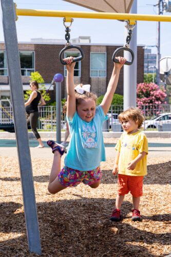 A girl holding onto overhead rings at the Jennie Dean Park playground in Arlington, VA, building upper body strength and coordination, installed by Sparks at Play using LSI equipment.