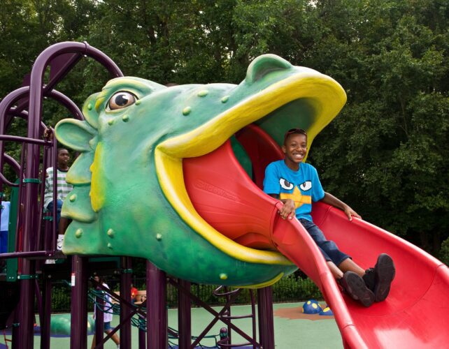 A child slides down a custom red dragon tongue slide, emerging from the open mouth of a giant medieval dragon play structure at South Bowie Community Center in Bowie, MD.