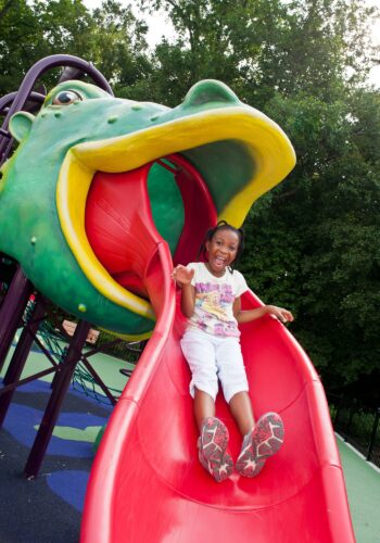 A child slides down a custom red dragon tongue slide, emerging from the open mouth of a giant medieval dragon play structure at South Bowie Community Center in Bowie, MD.
